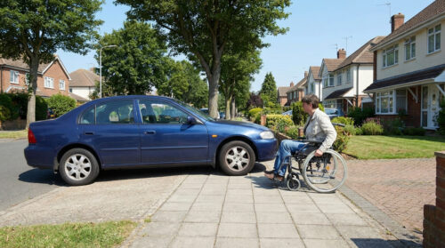 Une personne en fauteuil roulant bloquée par une voiture bleue garée sur un trottoir résidentiel, empêchant l'accès.