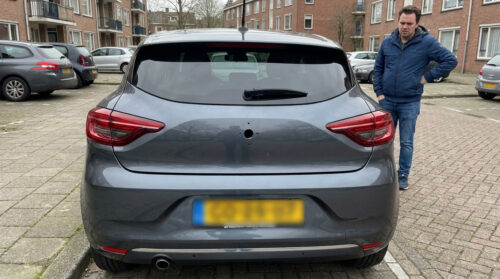 Une Renault Clio grise avec un trou béant sur le coffre là où se trouvait la caméra de recul, un homme regarde, dépité.