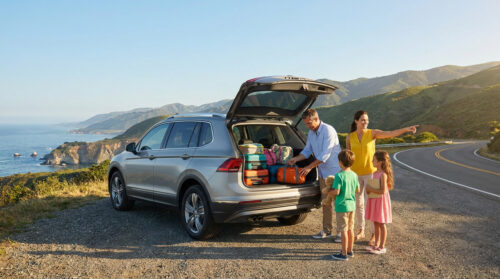 Une famille range ses bagages dans le coffre d'un SUV au bord d'une route côtière ensoleillée.
