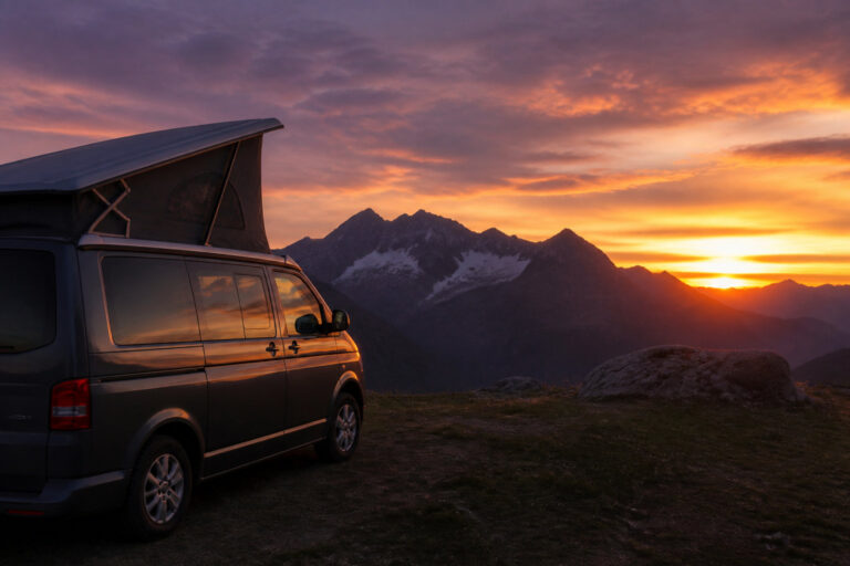 Un van aménagé avec toit relevable stationné sur un sommet montagneux devant un coucher de soleil orangé.