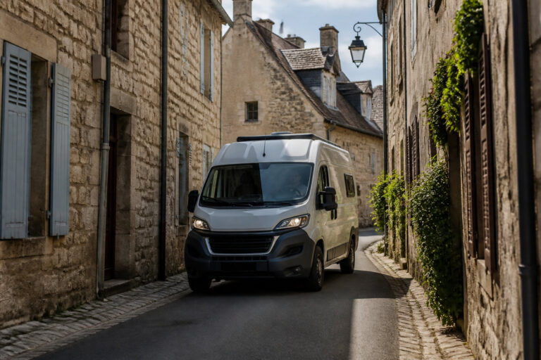 Un van aménagé blanc circule dans une étroite rue pavée entre des maisons en pierre dans un village français.