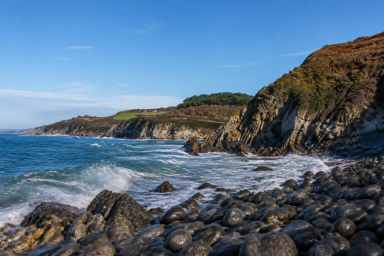 Paysage côtier rocheux avec des vagues s'écrasant sur une plage de galets sous un ciel bleu ensoleillé.