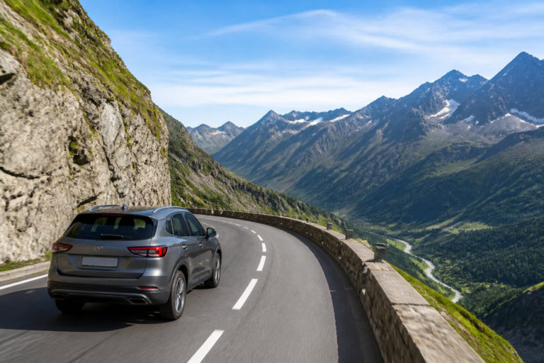 Une voiture grise roule sur une route de montagne sinueuse avec des sommets enneigés en arrière-plan.