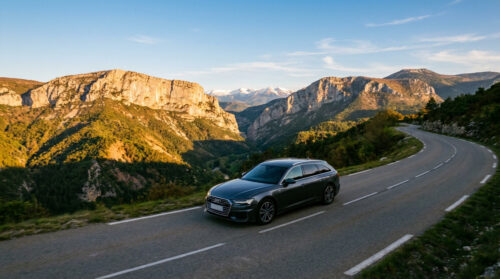 Une voiture Audi break grise roule sur une route sinueuse en montagne au coucher du soleil.