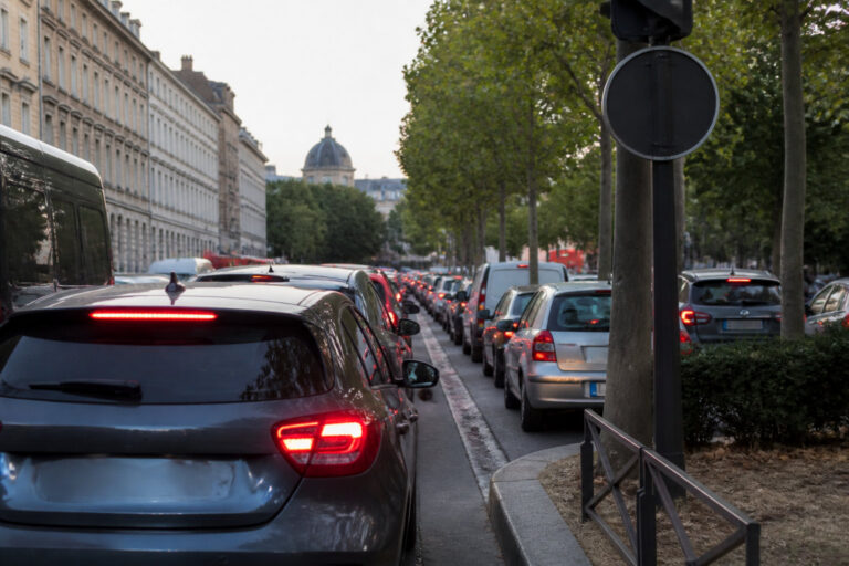 Une longue file de voitures bloquées dans un embouteillage sur une avenue bordée d'arbres avec des bâtiments anciens.